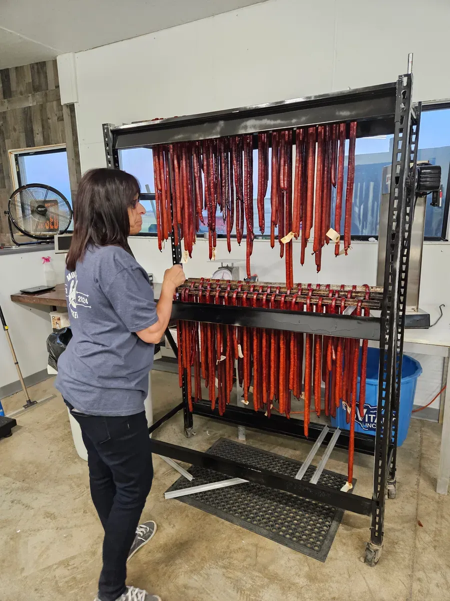 Mia Larson with racks of smoked meat sticks hanging to cool at H&M Custom Meat, Creston WA